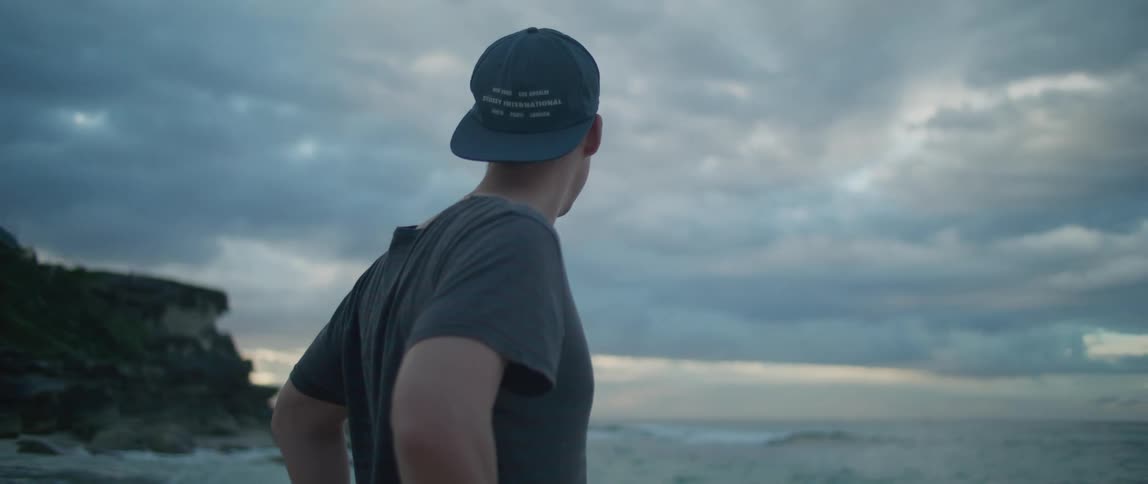 Man wearing a hat on backwards stands near a hill and looks out at the ocean