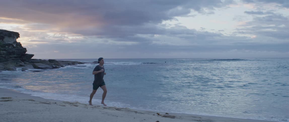 Man with a cap t shirt and shorts runs along the white sand beach next to the ocean at dusk