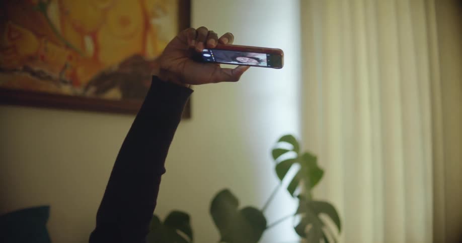 Young woman laying in bed talks on a video call on her phone inside a dimly lit room