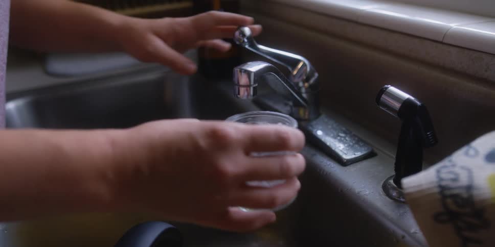 Woman fills glass from sink faucet before watering plants in window
