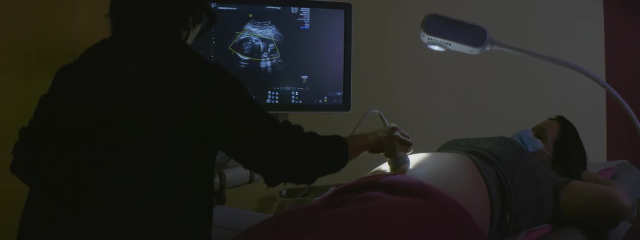 A nurse uses an ultrasound machine to take a sonogram of a pregnant womans belly in a dark examination room