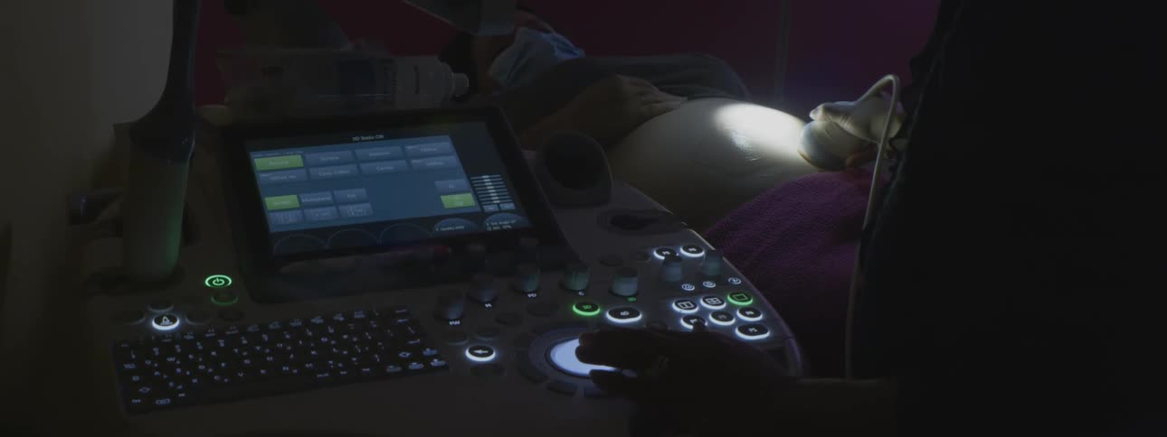 A pregnant woman gets an ultrasound in a dark exam room with light glowing from the machine and on her belly