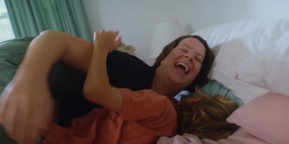 A woman a boy and a girl in a bedroom playing on a bed with a window in the background