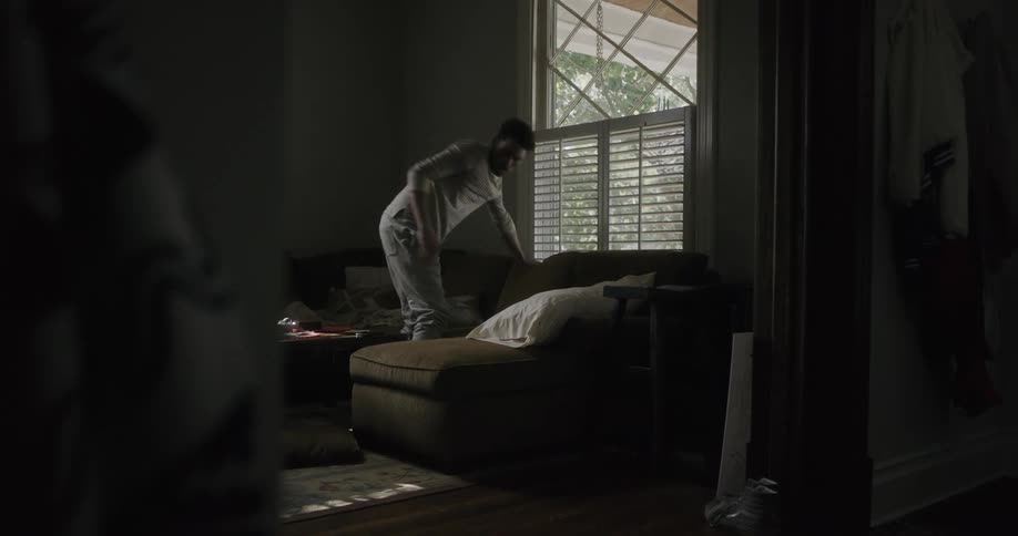Man gets up from the sofa and looks out a window then walks to another room and peers through the blinds in a door