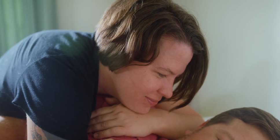 Woman smiles and hugs a man that is laying down with his head on a pillow