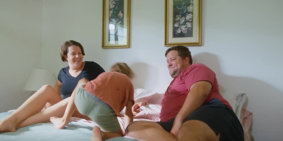 A boy runs through the corridor of a house into the bedroom of his parents where he begins to jump on the bed as they watch