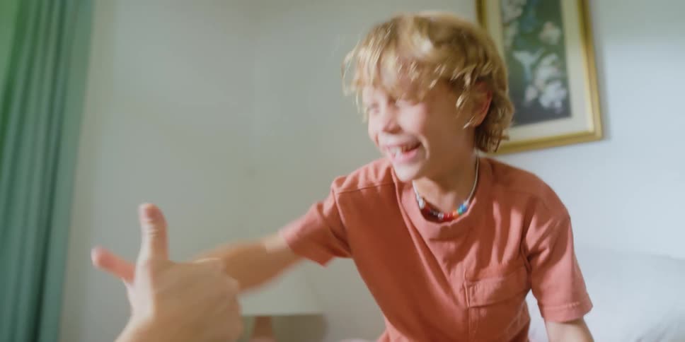 A boy plays thumb war with another person while sitting on the bed and laughs