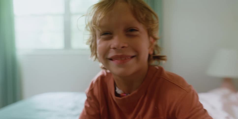 A young boy with blonde hair sits on a bed smiling for the camera before playfully falling backwards onto the bed