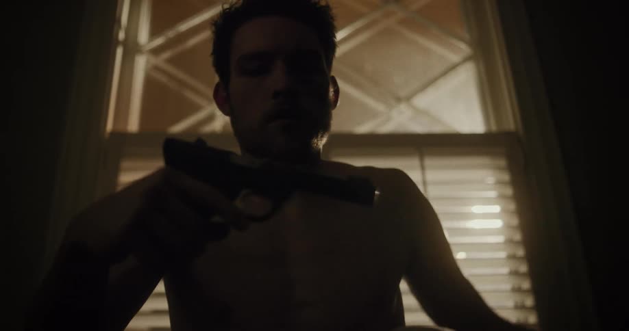 A man plays around with a black gun while sitting indoors in front of a window at night