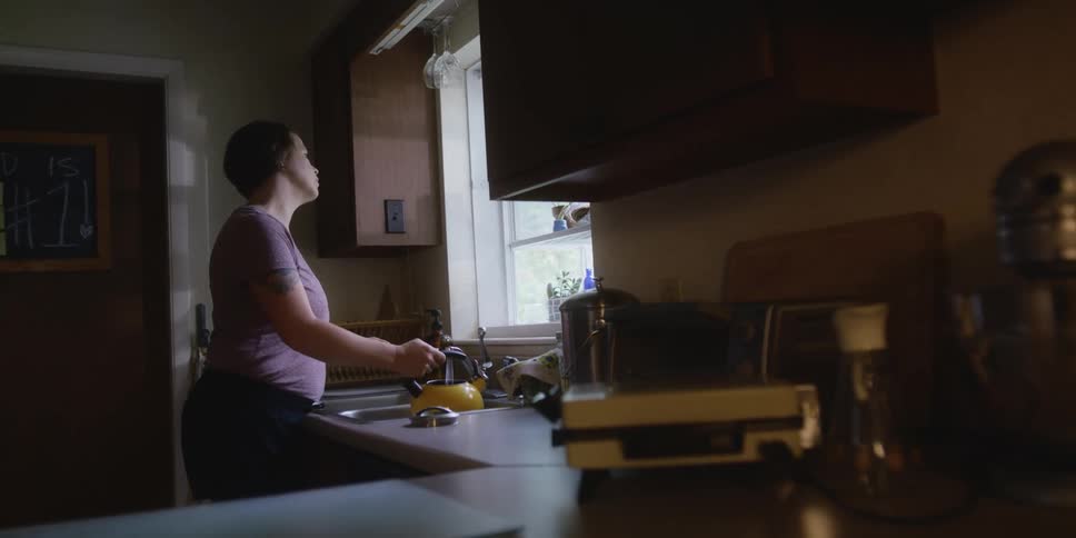 A woman stands at the kitchen sink filling up a tea pot from the faucet