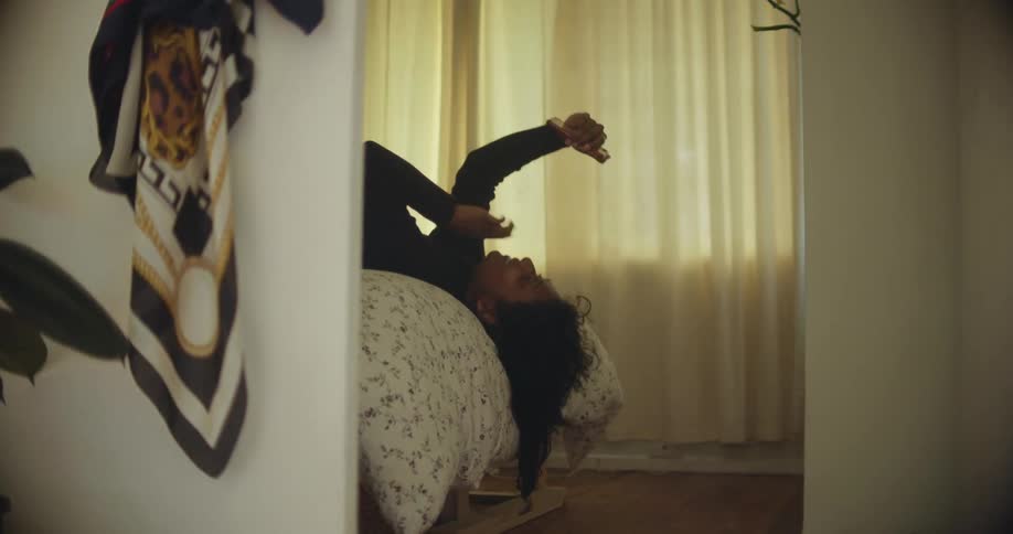 A young woman makes gestures with her hand as she participates in a video chat using her smartphone while lying on her back in bed