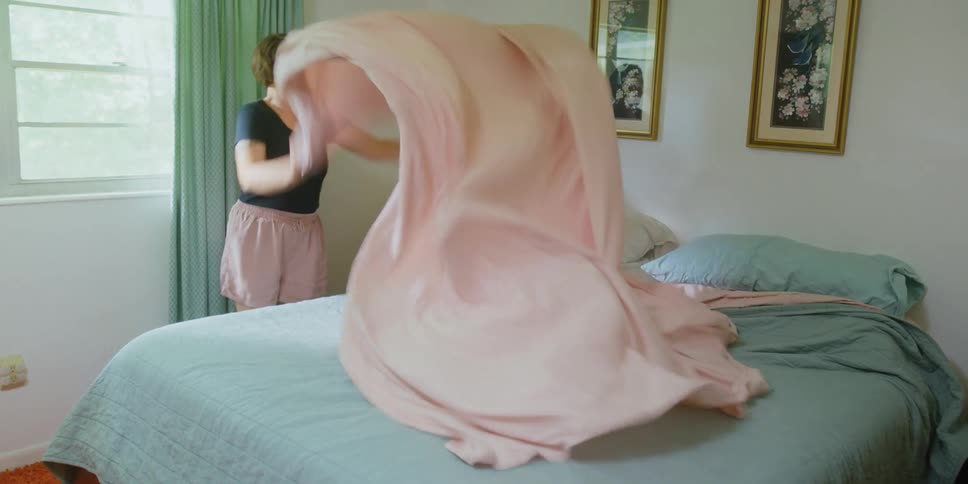 A woman throws a sheet on the bed playing with a young child laying on it