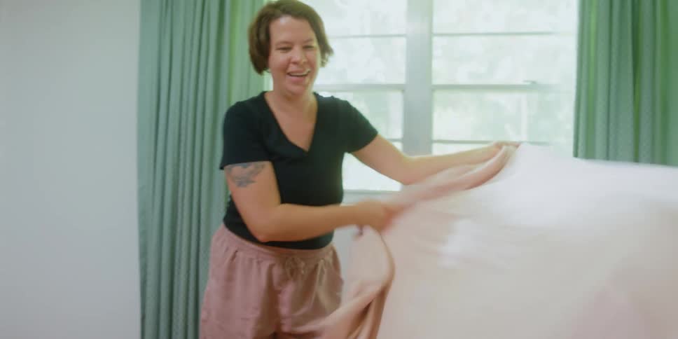 A woman throws a bed sheet over a pair of children lying on a bed before quickly pulling it off as the children laugh