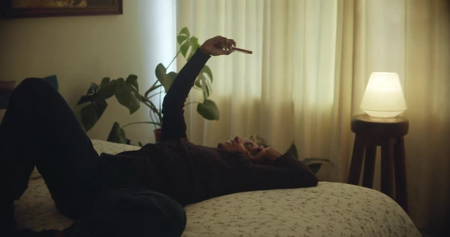 A woman waves at the camera of her smartphone while participating in a video chat while lying on her back in bed