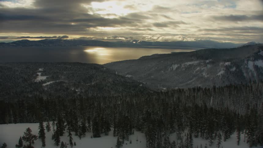 Aerial view of snowy mountains with forest next to lake as the sun shines through clouds