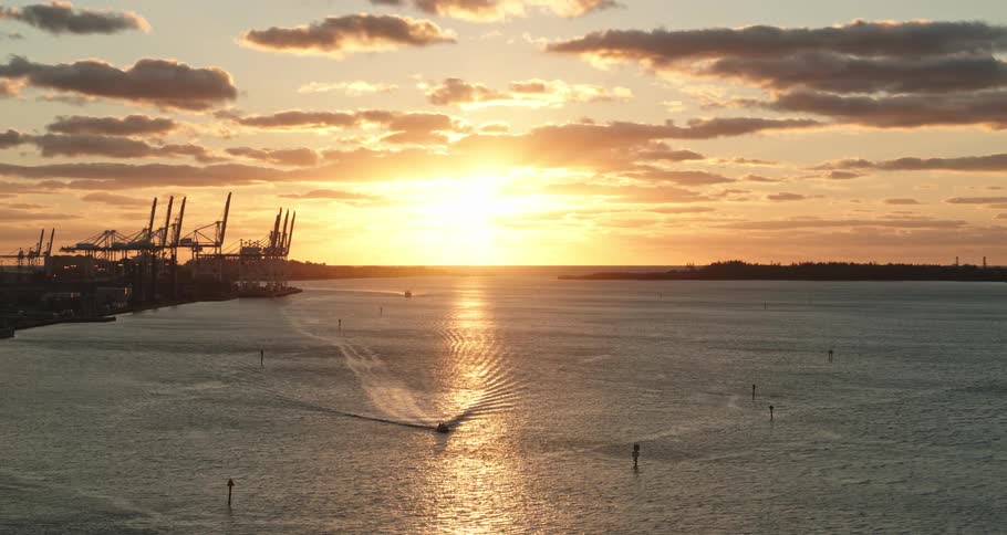 Sun shines from horizon turning sky red as boats move through water near shore docks and construction cranes