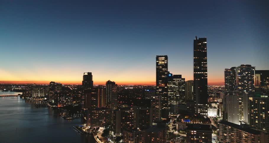 City lit by building lights stretches from water reflecting buildings to red tinted sky on horizon