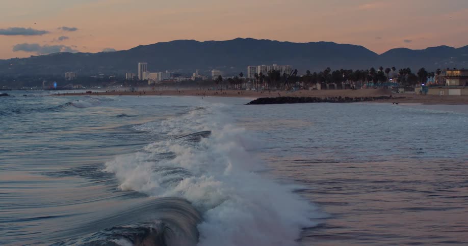 Ocean waves breaks against the beach with mountains and buildings in the background at dusk