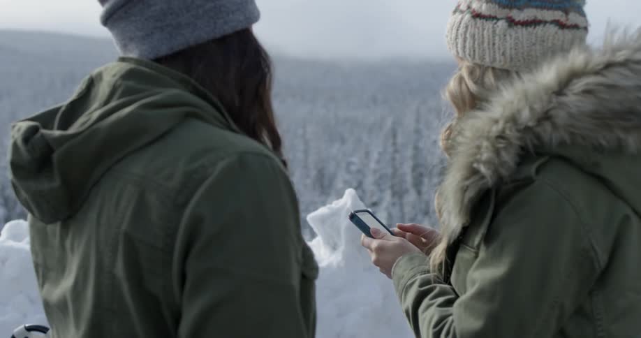 Two women stand on a cliff with a snow covered forest below