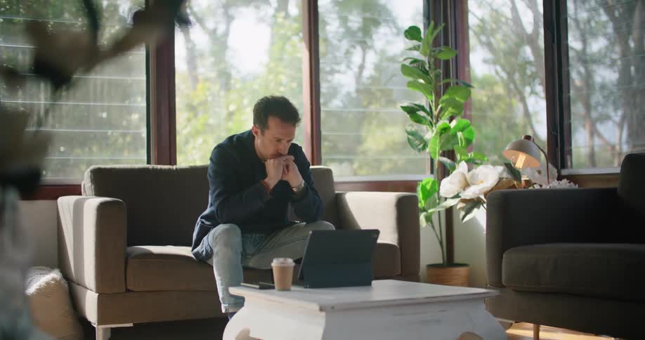 A man sits on a couch at home with a coffee and puts his chin in his hands as he looks at his laptop