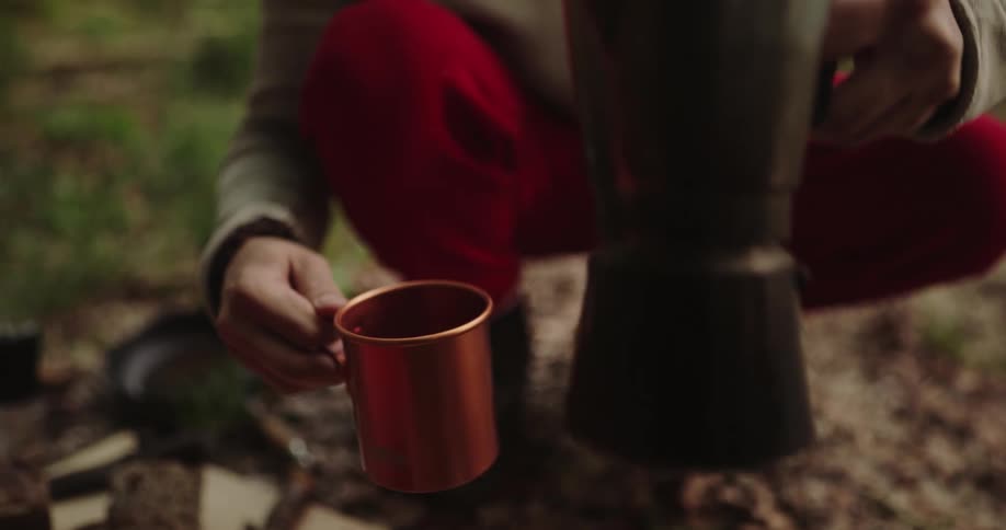 Person with red pants and beige sweater pours coffee into copper cup in the forest