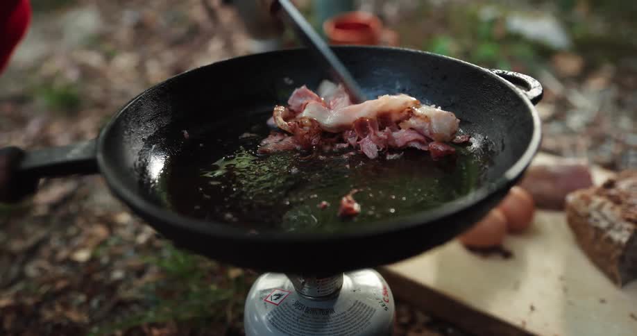 A man cooking bacon and eggs outdoors in a cast iron pan over a small heater with a gas tank