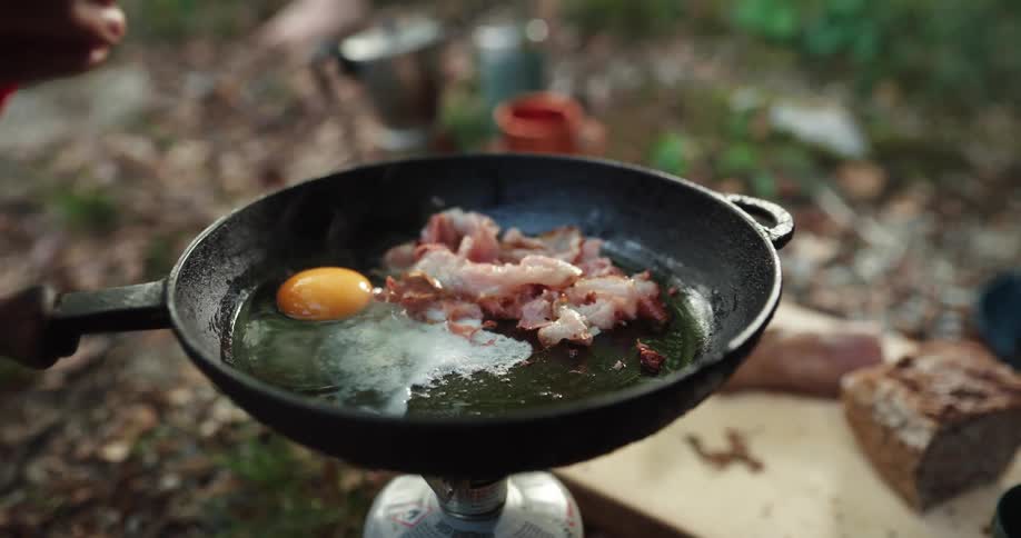 A person cooking outdoors cracks eggs in to a pan with bacon