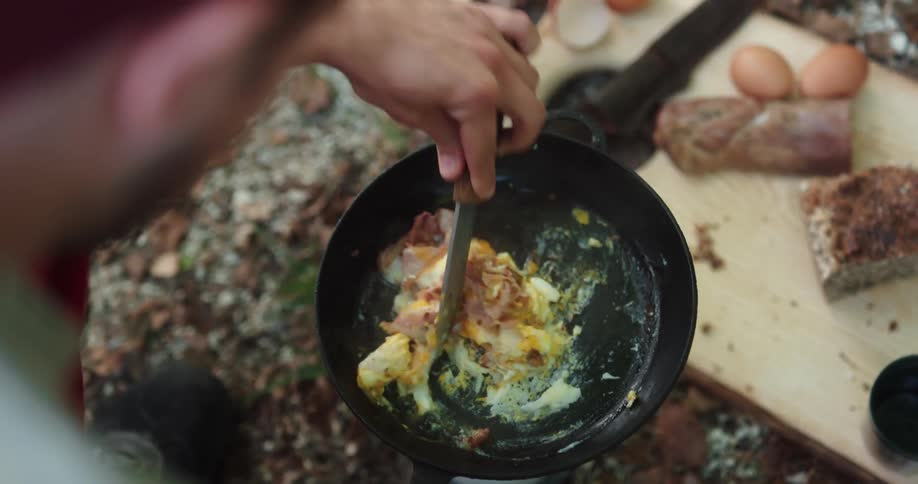 A man cooking bacon and eggs in a cast iron skillet outdoors in a forest over a small heater
