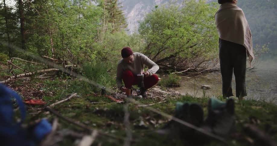 Woman wraps shawl around herself as she leaves tent to stand on shore near man preparing coffee