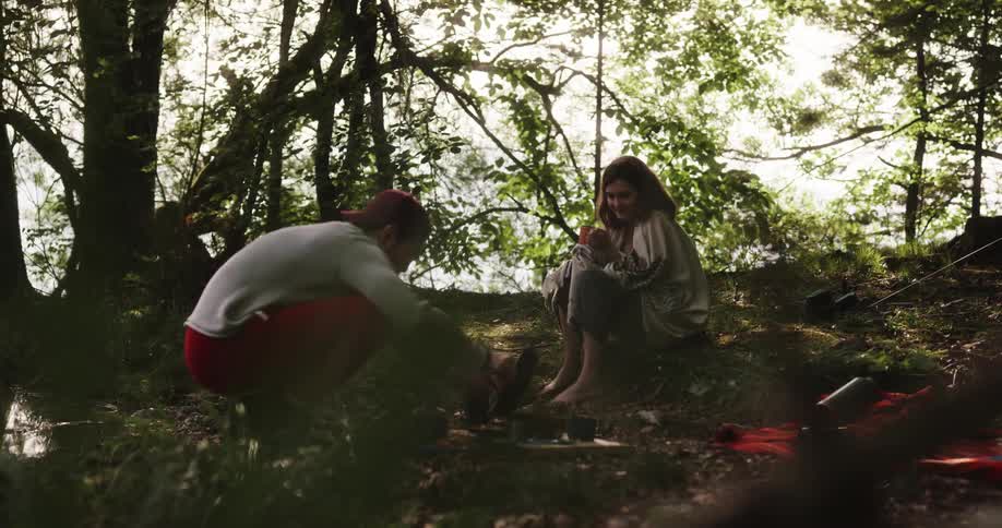 A man and woman are in the woods near a campfire that the man is cooking on
