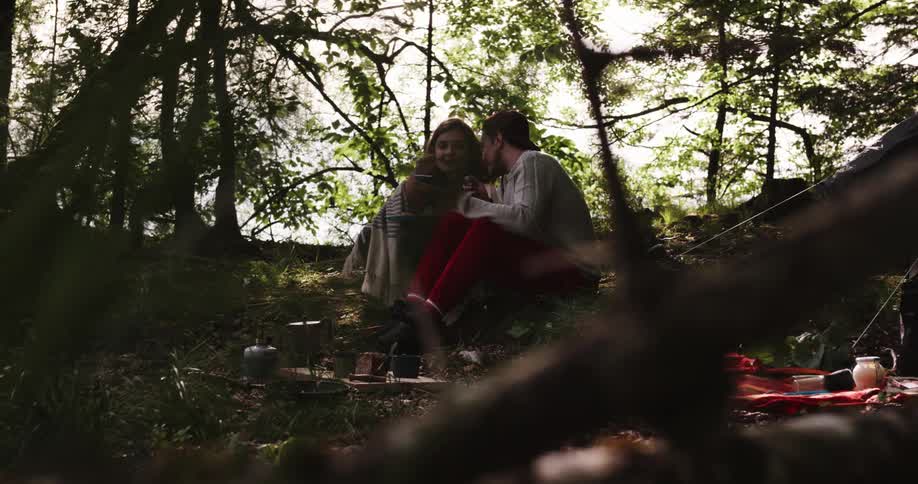 A male and female camper chat while they sit side by side on the ground and eat