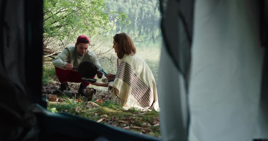 Couple shares meal cooked outdoors in field near tent with forest in background