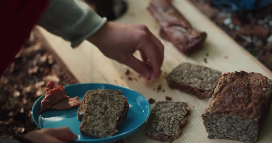 Person plates bread and meat from wooden chopping board set on ground before topping meal with sauce