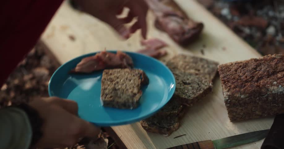 A camper cuts up a loaf of bread on a slab of wood and plates it with meat and sauce