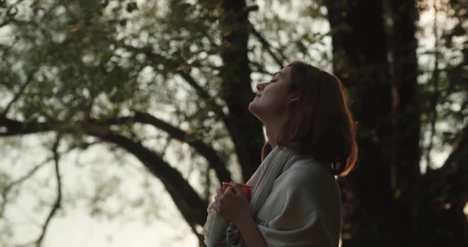 A woman standing outside next to a tree holding a coffee cup looking around and smiling