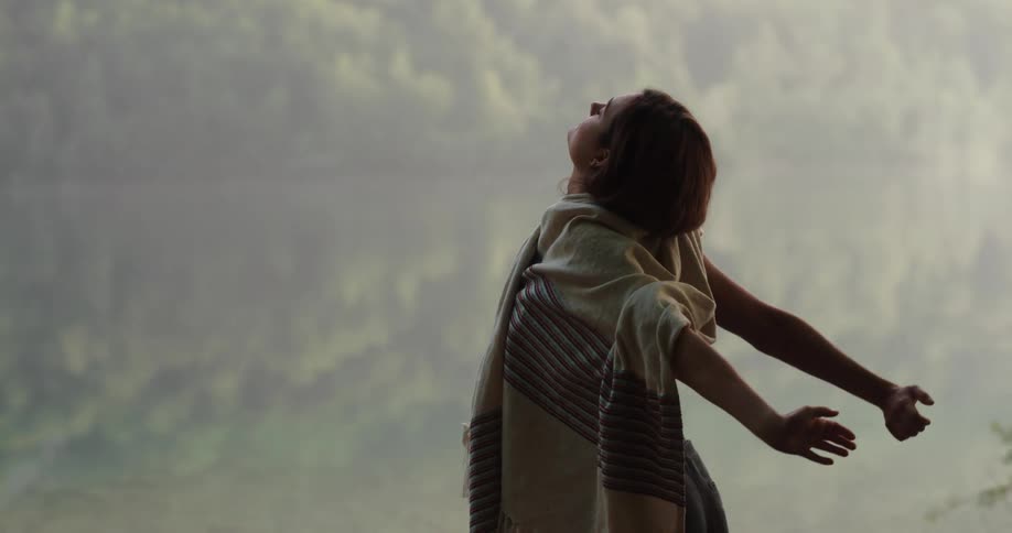 Woman wearing wrap and stretching stands near water reflecting forest on foggy day