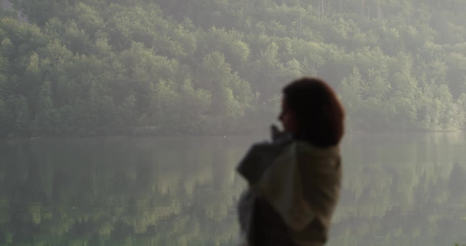 A woman adjusts the scarf around her shoulders as she stands near a lake