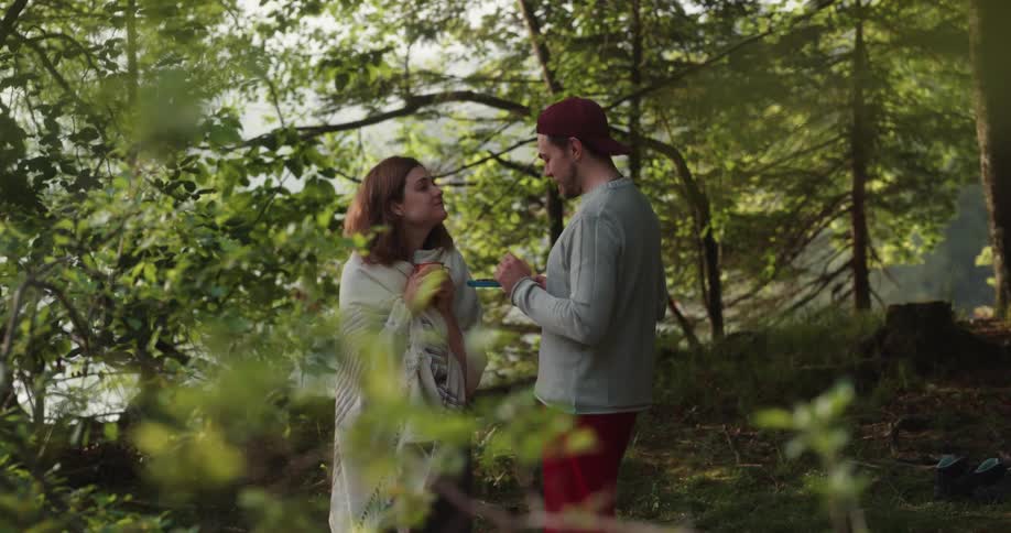 A man walks over to a woman standing in a forest covering herself with a blanket and feeds her from a plate then kisses her