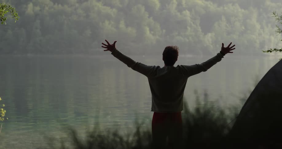 A man stands next to his tent by a lake and stretches out his arms