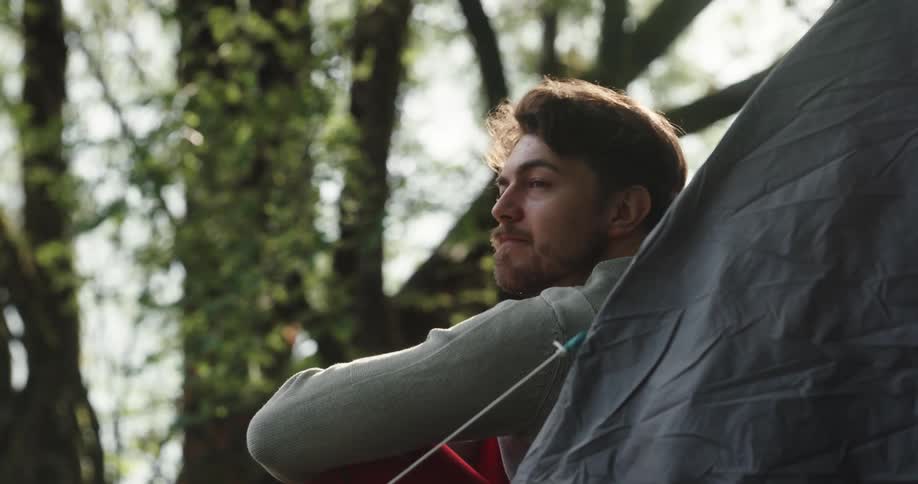Man with a beard a turquoise sweater and red pants sits down in front of a tent in the woods