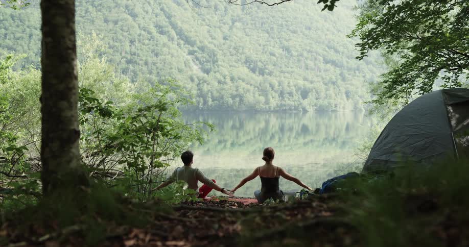 Man and woman do yoga before tent next to lake in the forest