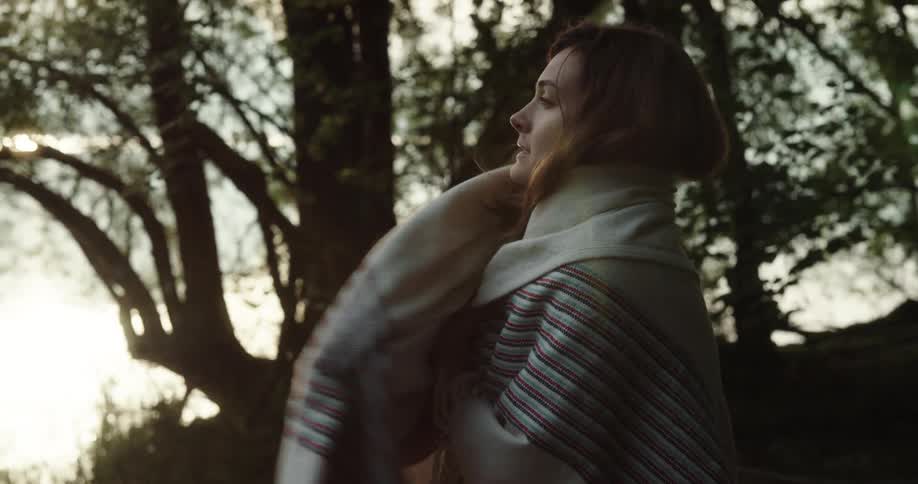 Woman feels cold and puts on shawl next to the lake with trees silhouetted against the sunset