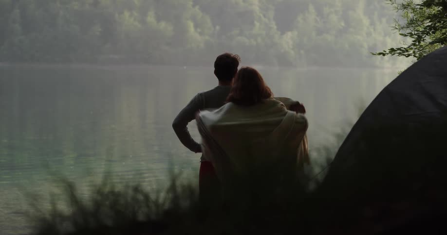 A man stands in front of a tent and looks out at the water and a woman emerges from the tent and goes up behind him to hug him
