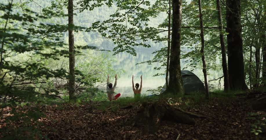 A man and woman practice yoga next to their tent on the shore of a lake in a forest during the day