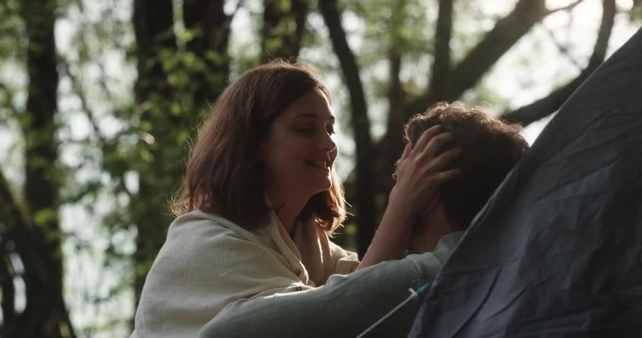 A woman kneels to kiss and run her fingers through the hair of a man sitting against a tent in a forest during the day