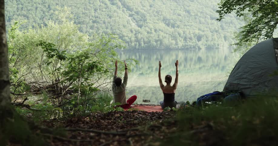 A couple sit facing a clear lake raising their arms in yoga stretches near their tent