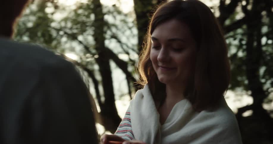 Man with a beard brings a copper cup of coffee to the woman with brown hair wearing a white shawl in the woods at sunset