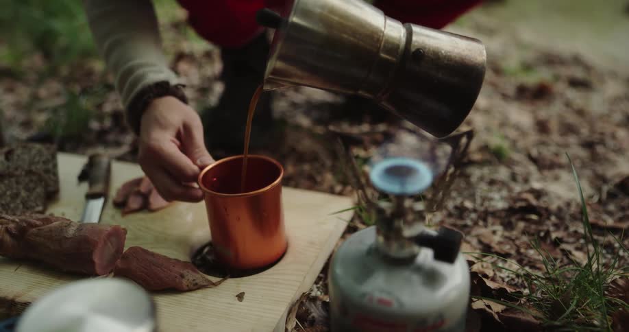 Man with red pants and boots puts coffee into copper cup in the woods