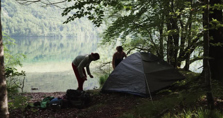 A man and a woman by a lake pitching a tent surrounded by trees