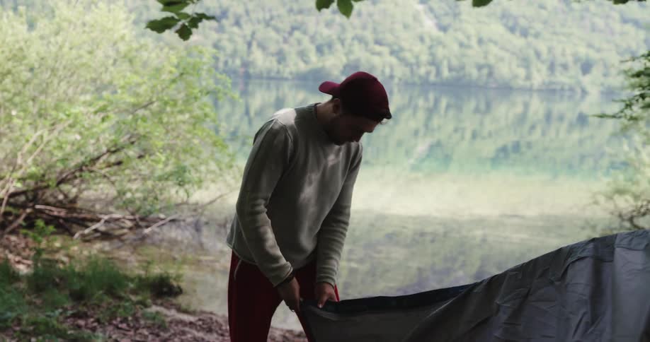 Man with red cap pants grey sweater and woman in black sleeveless shirt puts up tent next to lake in the woods